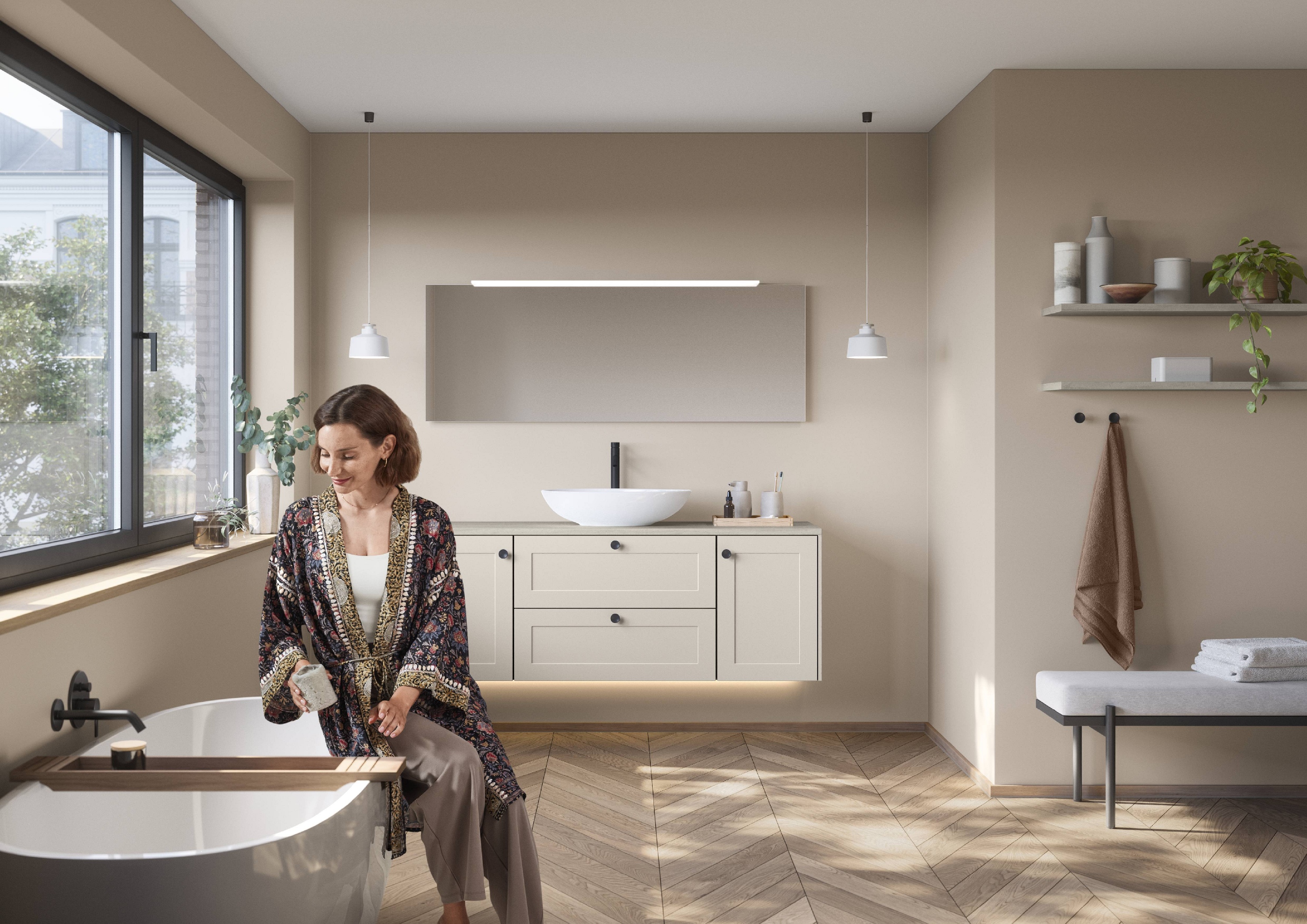 Badezimmer im modernen Landhausstil Fischgrätparkett weisse Waschschale auf cremeweissem Unterschrank mit Rahmenfronten und schwarzen Knaufgriffen Frau im feinen Morgenmantel sitzt lächelnd am Beckenrand ihrer freistehenden Badewanne und stellt Tiegel auf Wannenbrücke